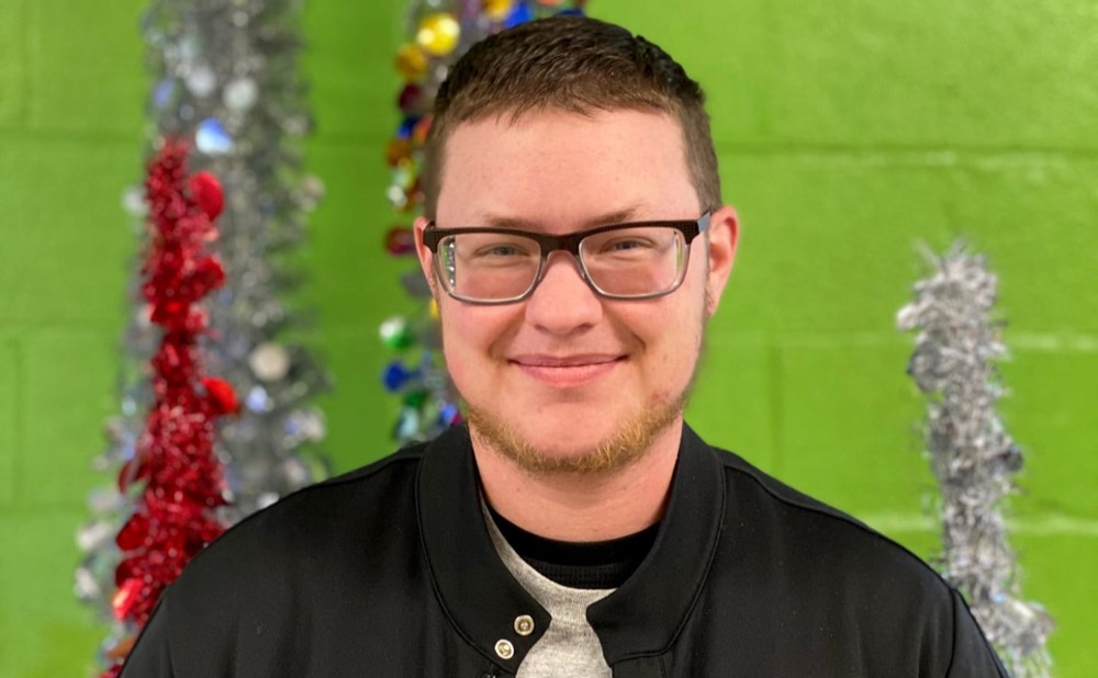 Portrait photograph of Dustin Parker, a young man in glasses and a black jacket, smiling in front of a green wall with tinsel decorations.