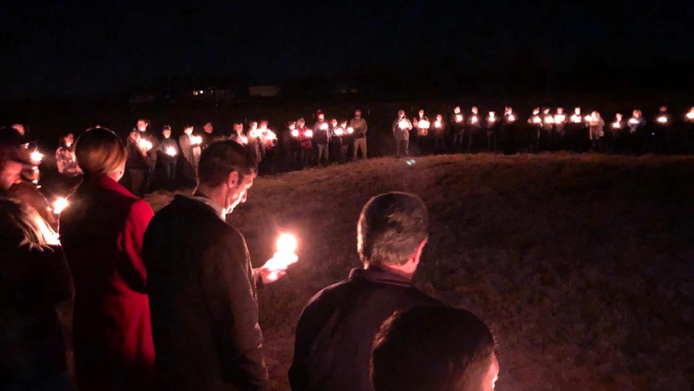 Night-time photograph of a large crowd standing in a field holding lit candles.